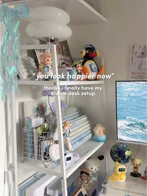 A well-lit, cozy corner of a room featuring a white shelf unit filled with various decorative items. The composition includes a prominent lamp, a collection of stylized figurines, books, and stationery items. A large monitor displaying blue water is present on the right. Layered text overlays are in the foreground, with additional text conveying what the person is grateful for.