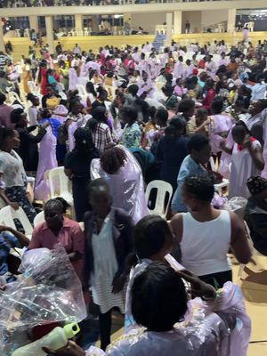 A large crowd of people, predominantly women, are gathered inside a well-lit hall. Many are wearing light-colored clothing. The image is taken from a slightly low angle, giving a sense of depth and scale to the gathering. Chairs are arranged, and the focus appears to be on the activity.