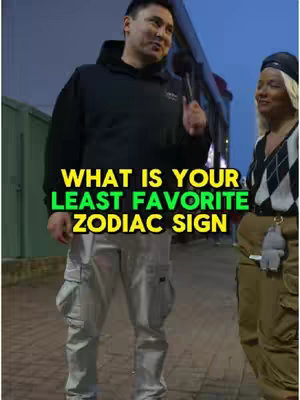 A street-level shot depicting a man holding a microphone addressing questions to people. The overlaying text on the image asks, "WHAT IS YOUR LEAST FAVORITE ZODIAC SIGN." The man is wearing a black hoodie and cargo pants. A woman stands to the right wearing a black cap and looking to her left. The image has a nighttime setting.