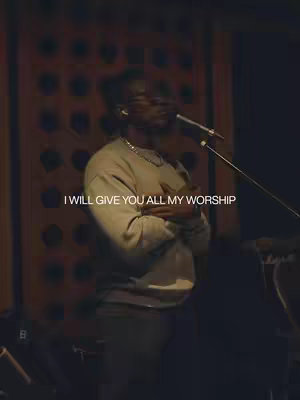 The image shows a man in a studio setting. He's wearing a light-colored sweatshirt, a silver chain, and is holding a microphone. The background shows a wood panel with a dark tone and there is a microphone stand. Text overlayed on the image reads, "I WILL GIVE YOU ALL MY WORSHIP."