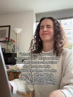 A woman with curly hair is looking towards the left side of the frame while smiling. She is wearing a white sweater and is indoors. The background includes a window, a lamp, part of a desk and a picture frame.