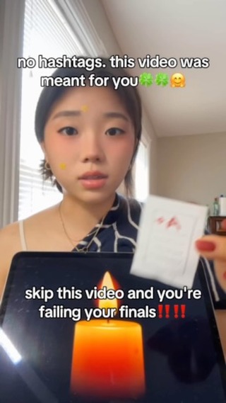 A young woman with a pale complexion is centered in the frame, looking directly at the camera. She has dark hair and is wearing minimal makeup. Her cheeks appear lightly flushed. A yellow sticker is on her forehead and another on her cheek. She's wearing a white camisole under a navy patterned top. She holds a white card, a piece of paper, with red markings. A tablet displays a glowing candle graphic in the background.