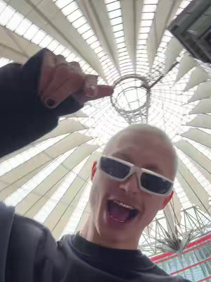 A selfie-style cover image, taken from a low angle, looking up towards a complex white and clear roof structure. A person with very pale skin, light hair, and sunglasses is in the foreground, with the person's hand raised, pointing upwards. The person's mouth is open, and the composition fills the frame with their face and the architectural background.