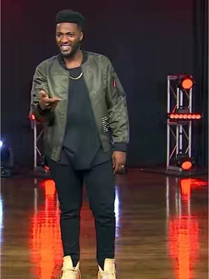 A man stands on a stage, gesturing with his hand. He wears a green bomber jacket and black pants.
