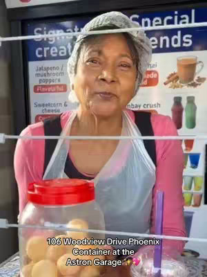 The cover image shows an older woman in a pink top and a hair net, smiling at the camera. She stands behind a glass display containing food, which has a jar with food items below her. In the background, a menu  is visible, along with bottles of beverages. The image is well-lit, focusing on the woman and her food stand.