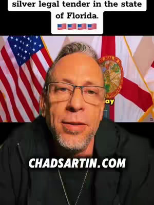 A man with glasses and a beard is centered in front of two flags. In the background are the American flag and the Florida state flag. The man is looking slightly up, and has a neutral expression. The overlay has the text "CHADSARTIN.COM"
