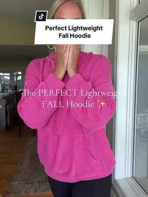 A woman stands indoors, likely in front of a window. She is wearing a vibrant, textured, pink hoodie and has her hands up near her face, possibly in a gesture of surprise or holding the garment up. The text overlay, in a white rectangle, states the hoodie's details. Additional text is on the image, and a TikTok logo in the top corner.