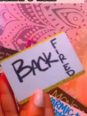 The image shows a hand holding a light blue card that says 'BACK FIRED'.  A pink patterned background is behind the card and hand grasping the card. Other card fragments that mention astrology are visible in the image.
