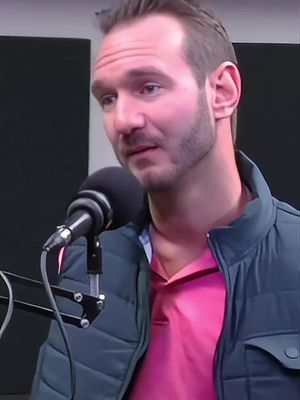 A medium shot features a man in a dark blue jacket, pink shirt, and a shaved head. Behind him, the studio-like backdrop fades into a gray shade. A black microphone sits centered in front of him.