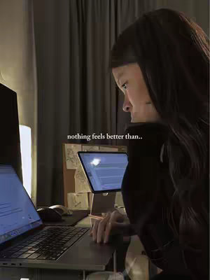 A woman is shown working on a laptop with a second screen and some notes on a corkboard visible in the background. She has dark hair, is wearing a black top, and is lit by the glow of her screens. Text overlayed on the image reads 'nothing feels better than...'.