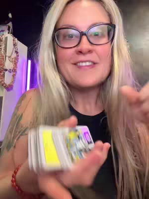 A woman with blonde hair and glasses is holding a deck of tarot cards, smiling at the camera. Her skin has a slight tan, and she wears a black top. The background is somewhat dark with a purple light on the left. There is a necklace and a red bracelet visible.