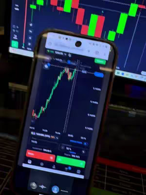 A smartphone displaying a financial chart is held up at an angle against a dark background. A computer monitor displaying similar charts is visible. The phone's screen shows a green upward trend line and red downward lines, indicating profit and loss, respectively. The overall composition focuses on the phone's screen with the trading charts as the central element.