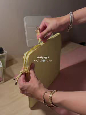 The video shows a person's hands opening a yellow pencil case on a wooden surface with a wall in the background.