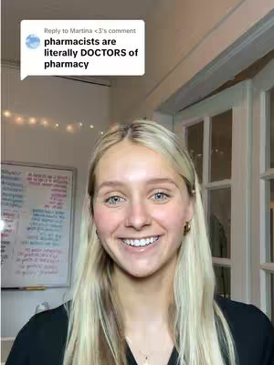 A young woman with blonde hair smiles directly at the camera. She is wearing a black top. Behind her is a white board with writing on it, fairy lights and white doors.