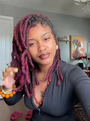 A woman with burgundy locs poses, gazing slightly down. She wears a black top and jewelry including a nose ring, necklace, and bracelets. Decor in the background includes wall art, and slippers on the floor.