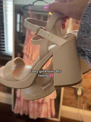 A hand holding a pair of beige platform sandals against a blurred background with a pink dress and a window.