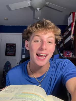 A young man with light skin and short, curly blonde hair is centered in the frame. He is wearing a blue t-shirt and a gold chain necklace. His mouth is open in a wide smile, and he appears to be speaking enthusiastically. Behind him is a blurry interior, including a ceiling fan, a door, and a partially visible poster.