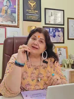 A woman in a floral top sits in a chair, facing the camera with a pen raised. Decorative items and framed pictures fill the background. She has a red and white dots on her forehead, big earrings, and a bracelet. A notebook with the words 'My Gratitude Journal' is visible on a table.