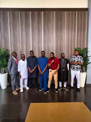A group of seven men of various ethnicities and attire are posing in a line. They stand in front of a wall with vertical wooden paneling that runs from floor to ceiling. To the sides are potted plants. The men are dressed in a range of styles, some formal and some more casual. One man is wearing a suit; others have button-down shirts and dress pants. The overall lighting is bright, and the composition suggests a group photo.