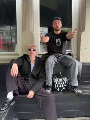 The image shows two men sitting outdoors. One is sitting on a black bag and the other is seated in front of him. The man in front, on the left, is wearing sunglasses and black clothes. The man in the rear is wearing a baseball cap, black t-shirt, and jeans. They are both gesturing with their hands.