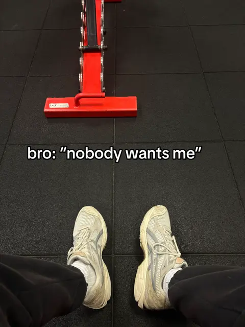The image shows a top-down view of a person's legs and feet in a gym. The person is wearing light-colored sneakers and dark pants. The floor is made of black, textured material. A red weight rack is visible in the background. Text is overlaid onto the image.