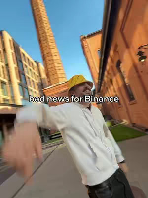 A man in a yellow bucket hat and white hoodie is standing on a paved walkway with buildings and a tall brick structure in the background. Text overlaid on the image reads bad news for Binance.