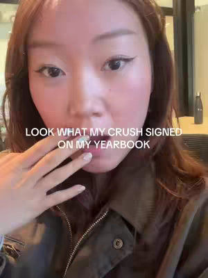 A selfie-style video featuring a young woman. She is shown from the chest up, her face centered in the frame. The lighting seems natural. She has long, brown hair, and is wearing what appears to be a dark jacket. Her hand is raised, partially obscuring her lower face and mouth.