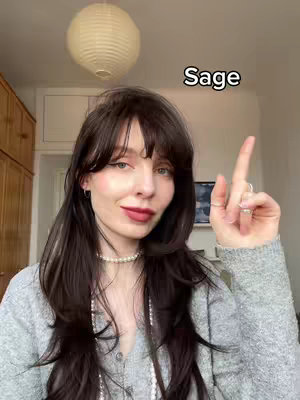 A woman with long dark hair and bangs points upwards in a room. She wears a gray sweater, and a pearl necklace. A text overlay at the top right corner states 'Sage'.
