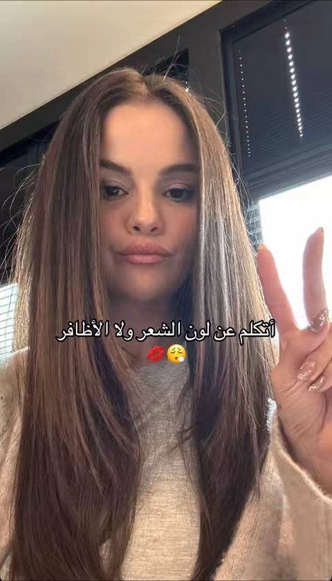 A selfie-style shot of a woman with shoulder-length brown hair, holding up her right hand in a peace sign. She has fair skin, and her lips are slightly pursed. The background is a blurred interior space with a black window. Text overlays in Arabic. Two emoji are also present.