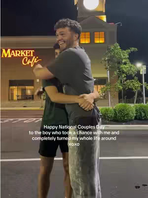 Two men embrace at night outside a building. One man is in the foreground, embracing the other from behind, with their faces close. The background includes a building with illuminated windows and a clock tower, and some greenery and parking lot.