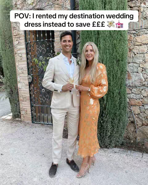 A well-lit photograph features a couple posing outdoors in front of a gate and hedge. The man, on the left, wears a light beige suit, a white shirt and black loafers. He holds a champagne flute and smiles at the camera. The woman, positioned to his right, wears a long-sleeved orange dress with gold floral designs and strappy high heels. Both are smiling. The background includes a stone wall and green foliage.