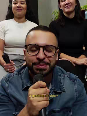 A medium shot featuring a man with glasses holding a microphone, positioned in front of two women. The man is centered, with the women slightly behind him, their faces and shoulders visible. The background is blurred, suggesting an indoor setting. The image has a warm, inviting feel due to the lighting and the expressions of the people.