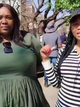 A selfie-style video shows two women outdoor in sunlight. The woman on the left is wearing a green dress and sunglasses, her arm rests in front of her. The woman on the right is holding a camera, with black and white striped shirt and a black baseball cap. Behind them are other people, trees.