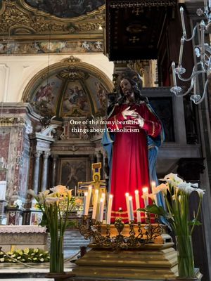 The cover image showcases the interior of a Catholic church, with a statue of Jesus prominently featured. The statue is depicted in a red robe. In front of the statue, there is an arrangement of candles. Floral arrangements, specifically white calla lilies, are visible on the left and right sides of the statue, adding a touch of elegance. The architectural background includes ornate details, arches, and paintings on the ceiling.