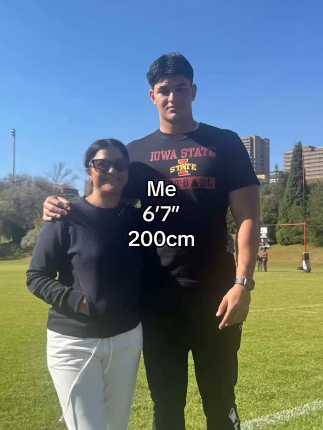A photo shows a tall man and a woman standing shoulder-to-shoulder on a sunny day. The taller man is on the right, wearing a black t-shirt with 'Iowa State' printed on it. The woman on the left wears dark sunglasses and a black sweater. The man has his arm around the woman's shoulder where there's a text overlay that reads 'me, 6'7', 200cm'. They stand on a green grassy field in front of a backdrop of trees and buildings.