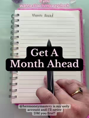 The image showcases a person's hand holding a pen over an open spiral-bound notebook. The notebook is open to a lined page where the words 'Month Ahead' are written at the top. A black banner across the page reads 'Get A Month Ahead' in white text. The person is wearing a gold ring with a black onyx stone.