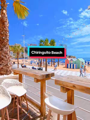 A vibrant image showcasing a beachfront scene in Benidorm. The frame is filled with a clear blue sky dotted with fluffy white clouds. In the foreground, a wooden bar area with white stools offers a view of the bustling beach. Palm trees and buildings line the promenade, while colorful beach huts and the sea create a lively backdrop. A text overlay in a bright color block advertises 'Chiringuito Beach,' setting the location. The composition is bright and inviting.