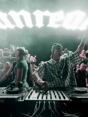 The cover image captures a live DJ performance at a concert or rave. The composition features the DJ central, facing the audience, with arms raised. The background includes a crowd partially visible behind the stage lights. The color palette is muted, with highlights from stage lighting. A text overlay in neon or bright lights.