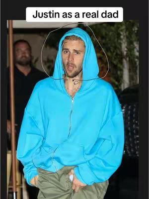 The image shows Justin Bieber in a light blue hoodie in a selfie-style shot. Above his head, there is the text'Justin as a real dad' in white font with a slightly blurred background. He appears to be outside, possibly in a city area, and a person is partially visible in the background to the left. The image is well-lit, and the colors are natural, with the blue hoodie offering a vibrant contrast to the background.