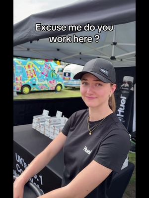 A young woman wearing a black cap and t-shirt with the Huel logo is smiling at the camera. Behind her is a Huel-branded truck and outdoor setting.