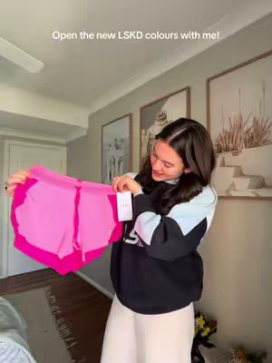 A woman is holding up a pair of pink athletic shorts, presenting them in front of her as if opening the packaging. She is smiling and wearing a black and light blue sweatshirt and light-colored pants, standing inside a well-lit room. The background includes framed artwork.