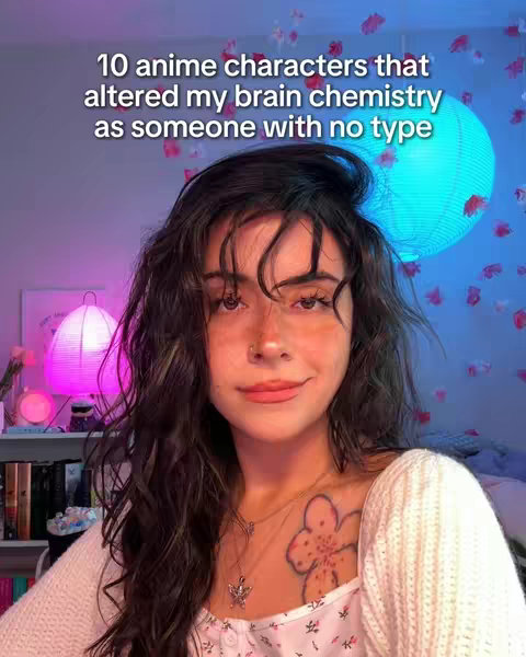 A young woman with long, dark curly hair smiles towards the camera. She wears a light-colored, open-front sweater over a floral top, revealing a tattoo on her chest. The background includes a shelf of books, glowing pink and blue orbs, and decorations.