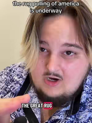 A person with light brown hair, wearing a blue and white patterned shirt, and dark suspenders, points to the bottom of the screen while speaking. The background is a light beige color and appears to be an office or personal space. Text overlays stating, “the rug pulling of america is underway” and “THE GREAT RUG” are clearly visible.