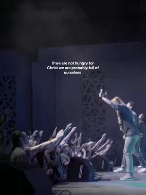 A stage is lit with blue light, and a man is on stage with his arm raised toward an eager audience. Those in the audience are reaching toward the man on the stage with their hands in the air.