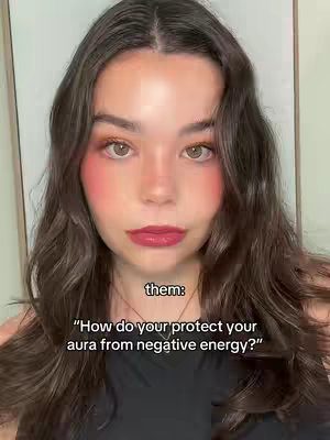 A close-up shot of a woman with long, wavy brown hair and makeup. She's looking directly at the camera. Text overlayed at the bottom says "them:" and a question about protecting one's aura.