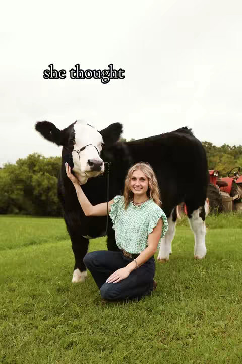 A young woman kneels on a green lawn, gently touching the face of a black and white cow wearing a pair of glasses. The woman smiles at the camera, and the background has a rural scenery with trees and some farming vehicles in the background, the sky is overcast.