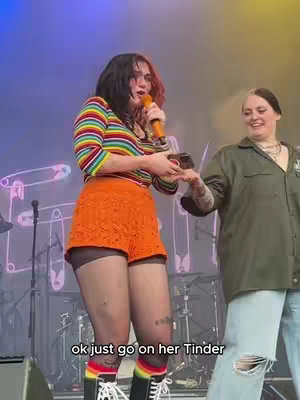 Two women are on a stage. One with a rainbow striped top and orange shorts is holding a microphone and a phone. The other is standing slightly behind her wearing a green shirt and blue jeans. There is a stage and drums in the background.