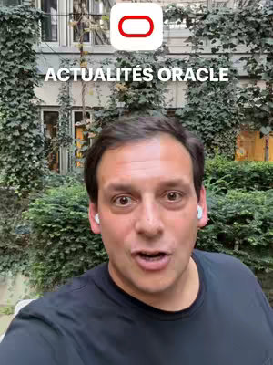 A man with brown hair is standing in front of a building with green bushes and vines, with an overlaid logo in the top center stating "ACTUALITÉS ORACLE". The man is wearing a black t-shirt and wireless earbuds. He's speaking, likely to camera. Light comes from the right side of the frame.