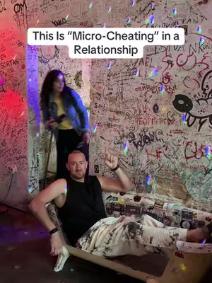A man in a black tank top is seated in a chair, pointing upwards. Behind him and the chair, a woman stands on a step. The background is a wall completely covered in graffiti with many different colors, with a disco ball shining lights on top.