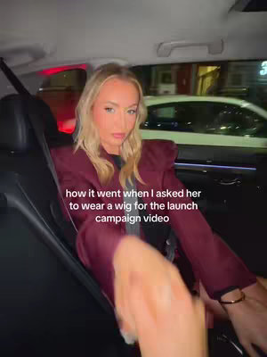 A woman in a car wearing a dark burgundy blazer stares directly at the camera. The text overlay reads, "how it went when I asked her to wear a wig for the launch campaign video."